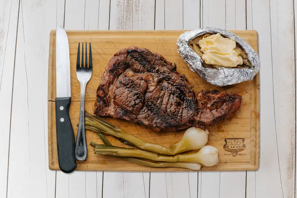 Rib eye asado servido en tabla con papa al horno, ejotes y cebollitas